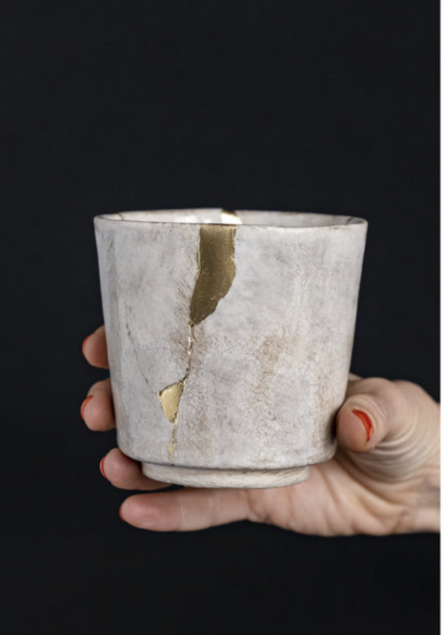 Woman's hand holding a light gray pottery cup that has been mended with gold in the Kintsugi technique.
